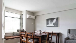 A dining area with a wooden table and chairs, a bench with cushions, a wall-mounted air conditioner, and a framed picture.