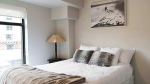 A neatly made bed with white bedding and a fur throw, a lamp on a bedside table, and a framed picture on the wall. - The Rainbow Suite Mountain and Village View (Whistler)