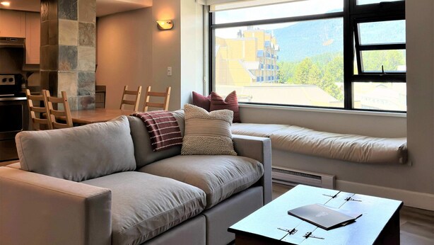 A modern living room with a sofa, a coffee table, a dining area with chairs, a fireplace, and a large window with a view. - The Rainbow Suite Mountain and Village View (Whistler)