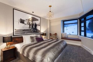 A bedroom with a large bed, a framed historical photo, a window with a view of a snowy landscape, and a bench by the window.