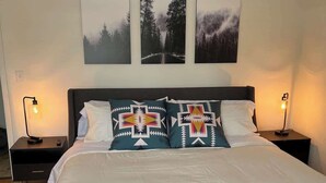Una cama bien hecha con ropa de cama blanca y dos almohadones decorativos, flanqueada por dos mesitas de noche con lámparas, en una habitación con techo inclinado y tres cuadros en la pared.