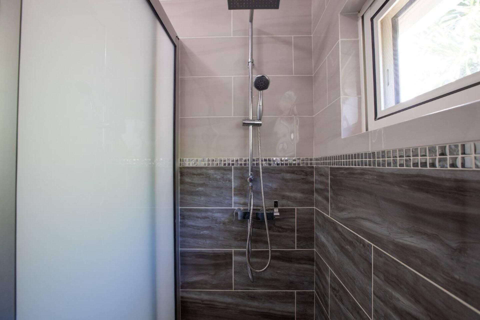 Ducha con cabezal de ducha manual y un cabezal fijo en el techo, paredes de azulejos y una ventana.