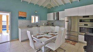 Cozinha moderna com armários brancos, eletrodomésticos de aço inoxidável e área de refeições com mesa e cadeiras brancas.