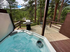 A hot tub on a deck with a grill and chairs, surrounded by trees.