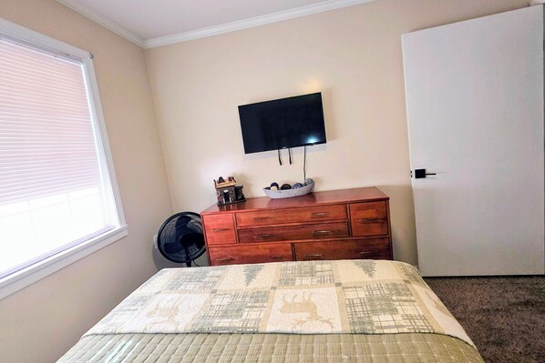 A bedroom with a bed, a dresser, a TV mounted on the wall, a fan, and a window with blinds.
