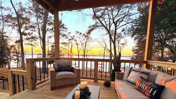 Una terraza de madera con sofá y sillón, una mesa con velas, y vistas al atardecer sobre un cuerpo de agua.