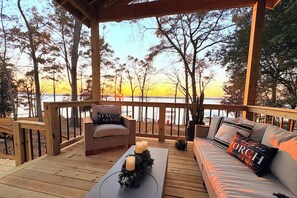 Une terrasse en bois avec un canapé et un fauteuil, une table avec des bougies et une vue sur le coucher de soleil au-dessus d’une étendue d’eau.