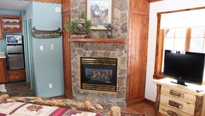 Una habitación acogedora con chimenea de piedra, un televisor sobre un soporte de madera, una cama con colcha y una zona de cocina con electrodomésticos.