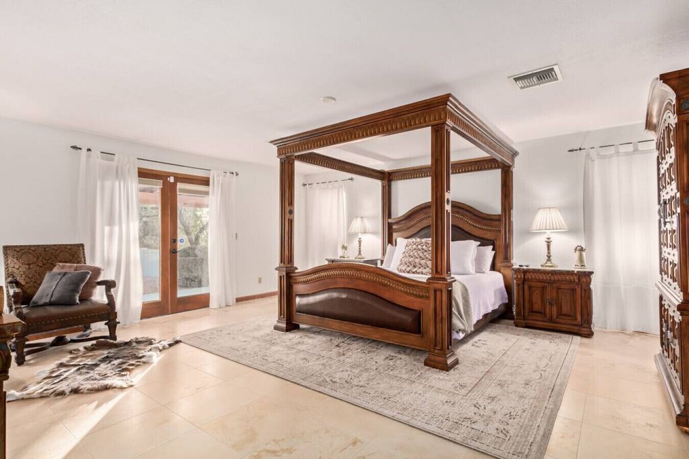 A four-poster bed with a wooden frame, a sitting area with a chair and a small table, and a large window with curtains.