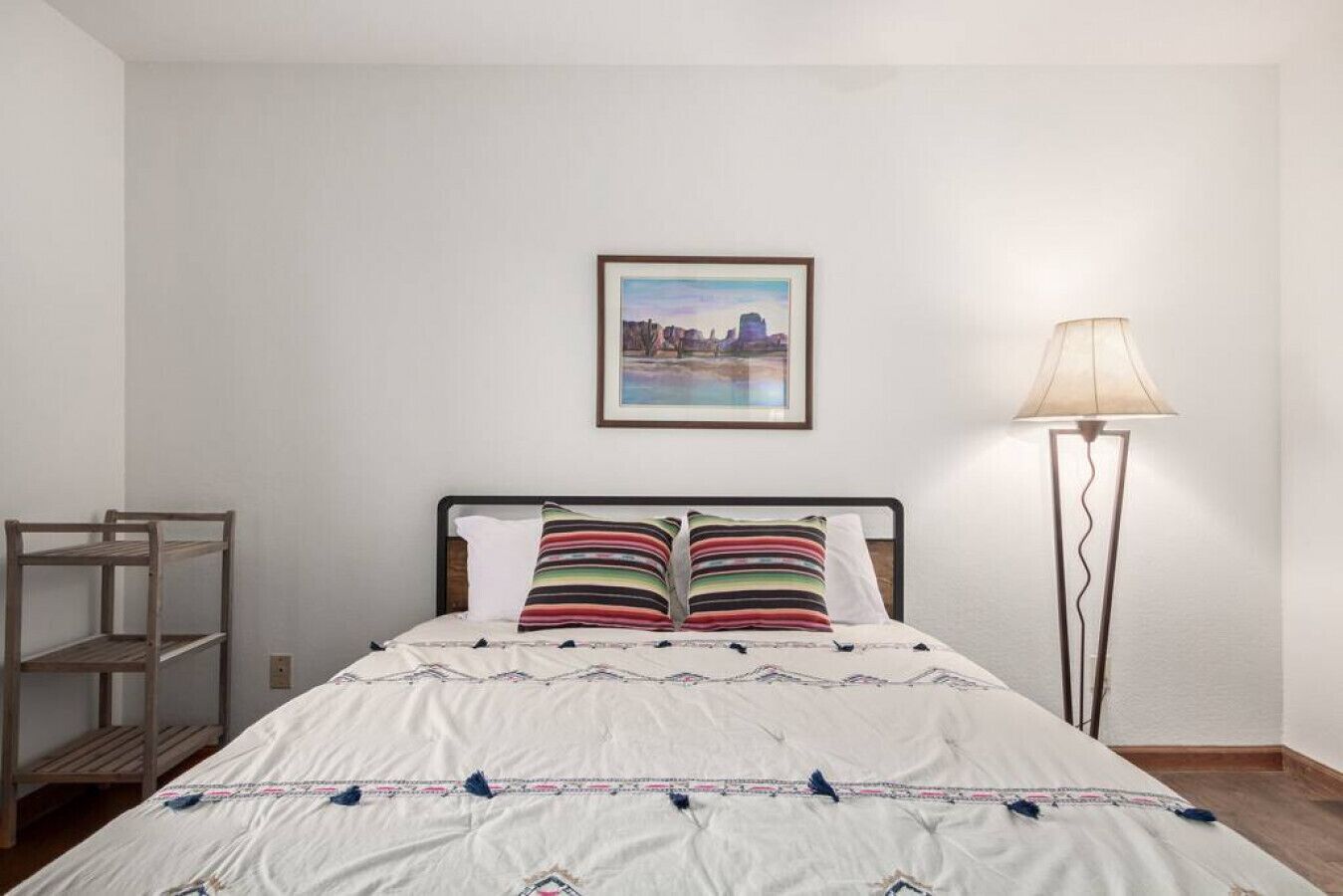 A bedroom with a bed, a framed picture on the wall, a floor lamp, and a wooden shelf.