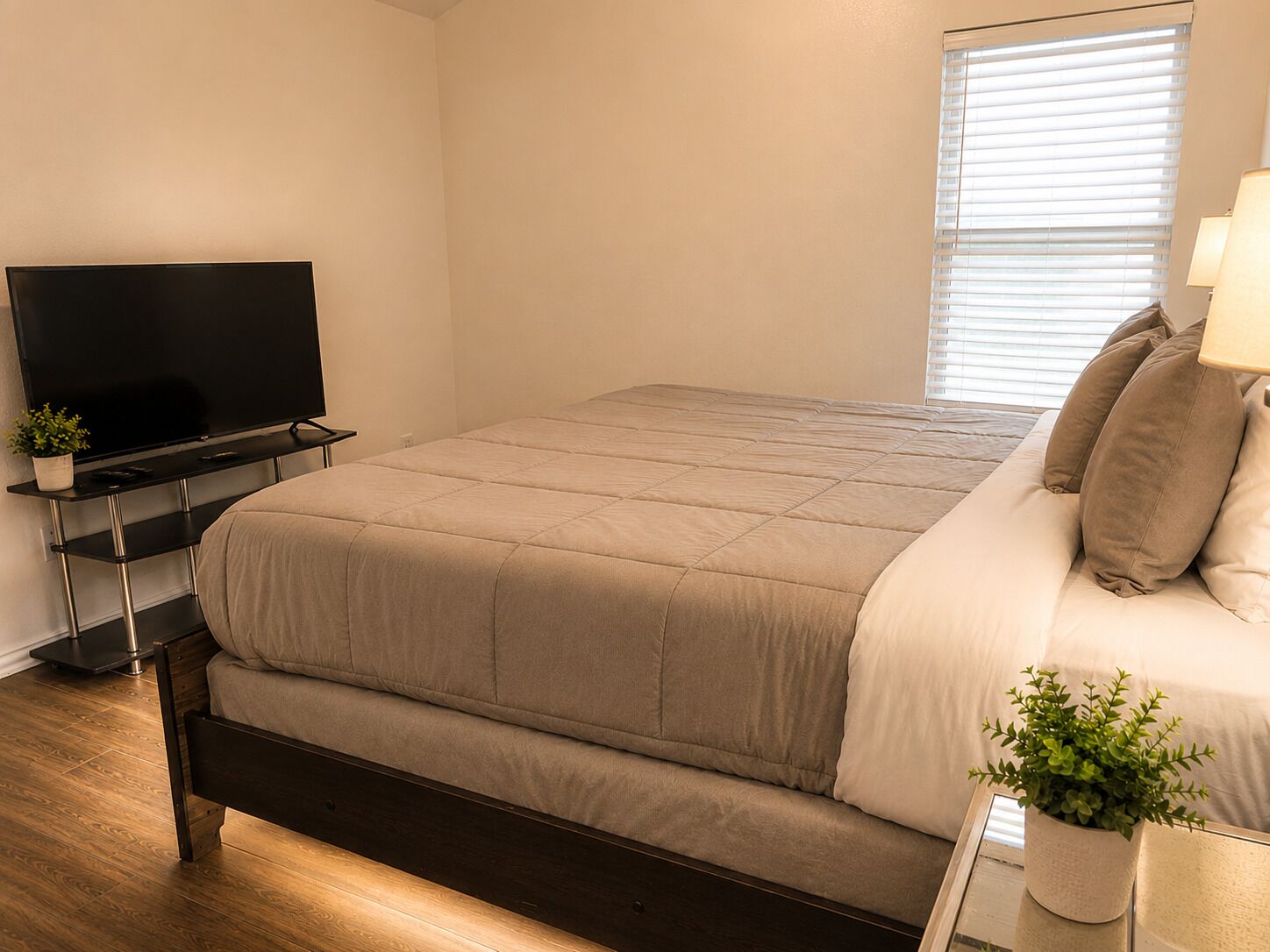 Ein Schlafzimmer mit einem Bett, einem Fernsehgerät auf einem Schrank, einem Fenster mit Jalousien und einem Nachttisch mit Lampe und Pflanze.