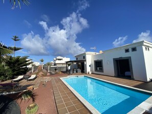 A swimming pool with a tiled deck, lounge chairs, and a white building with a garage. - Luxury villa with private heated pool (PLAYA BLANCA)