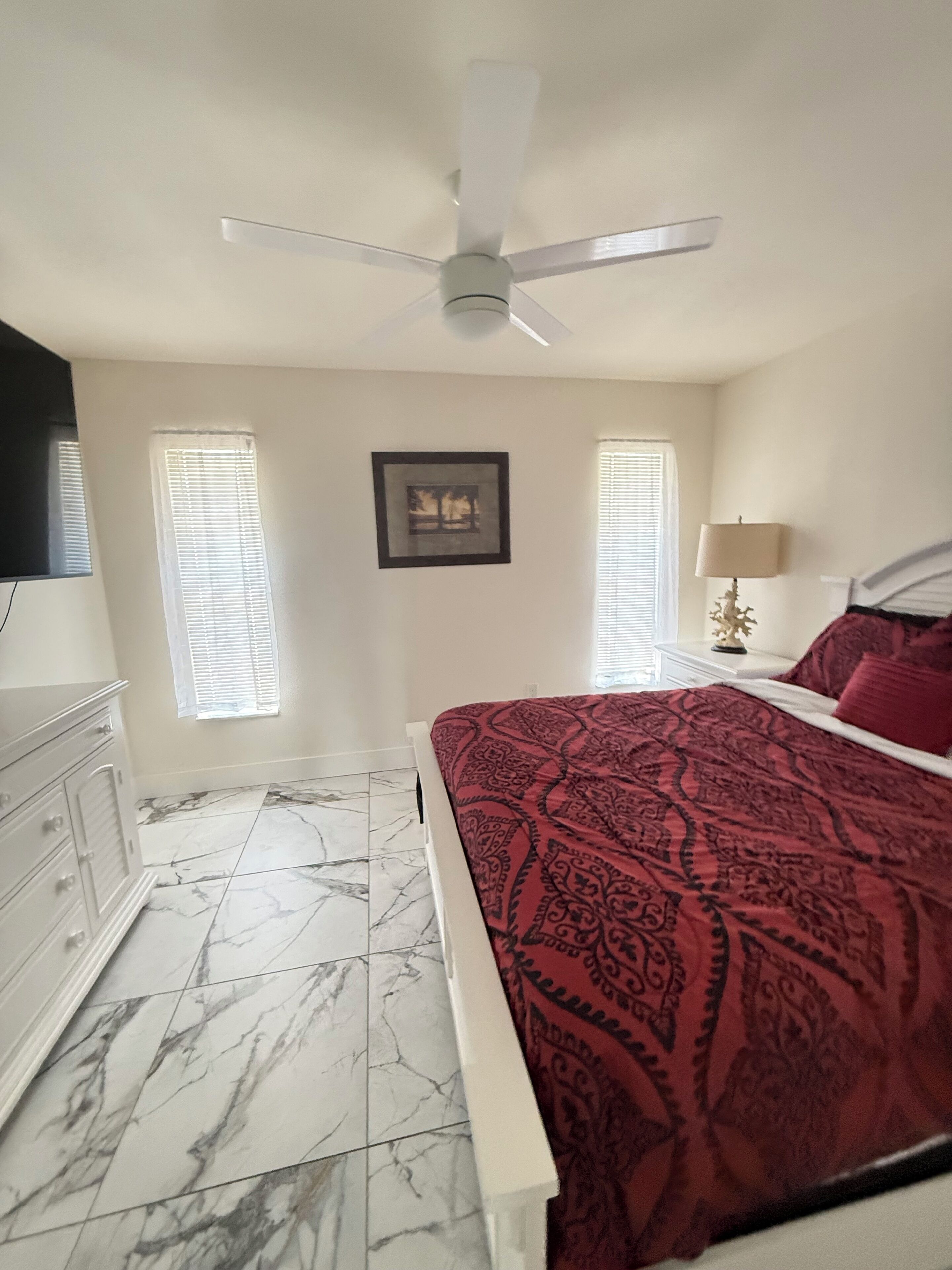 A bedroom with a bed, a ceiling fan, a nightstand with a lamp, and a dresser.