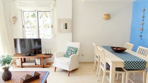 Ein Wohnzimmer mit Fernseher, einem weißen Sessel, einem Esstisch mit Stühlen und Blick auf Palmen durch das Fenster.