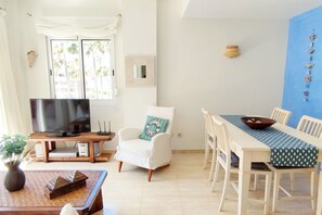 A living room with a TV, a white armchair, a dining table with chairs, and a view of palm trees through the window. - Apartament Flamingo by DENIA COSTA (Dénia)