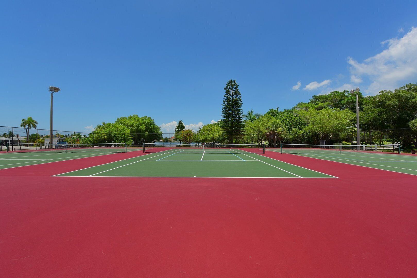 A tennis court with a red surface and green boundary lines, surrounded by trees and a clear sky.