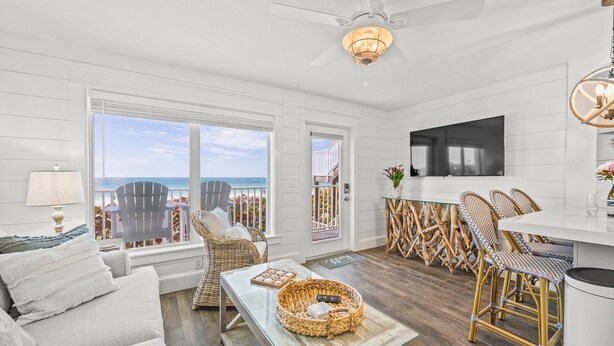 Ein geräumiges Wohnzimmer mit großem Fenster zum Strand, Korbmöbeln und einer modernen Küchenzeile.