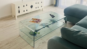 Ett modernt vardagsrum med ett soffbord i glas, en platt-TV som är monterad på väggen, ett vitt avlastningsbord och en blå soffa.