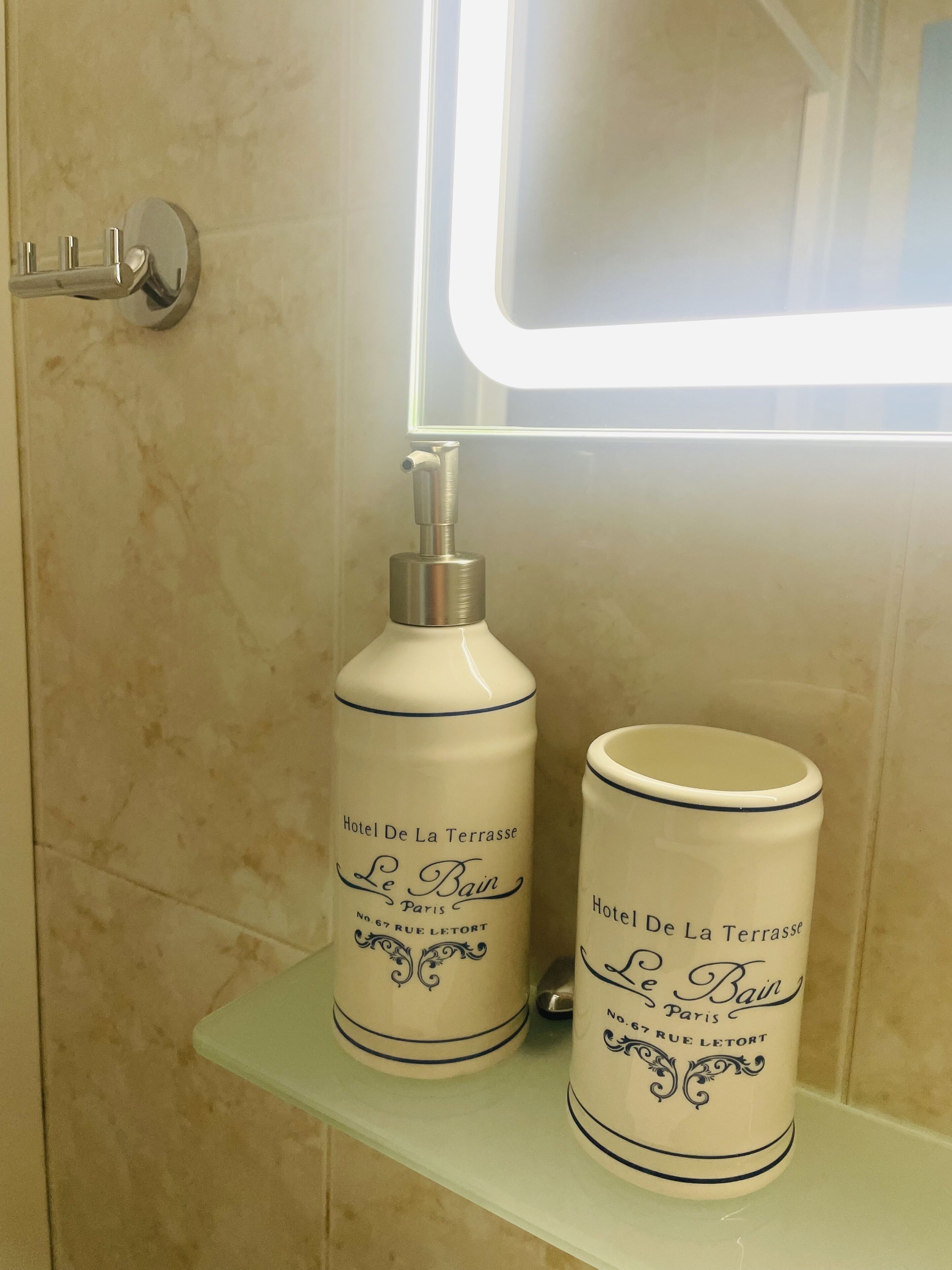 Une salle de bain avec un mur en marbre blanc, une douche en verre et deux récipients blancs portant l’inscription « Hôtel De La Terrasse Le Bain Paris », placés sur une étagère.