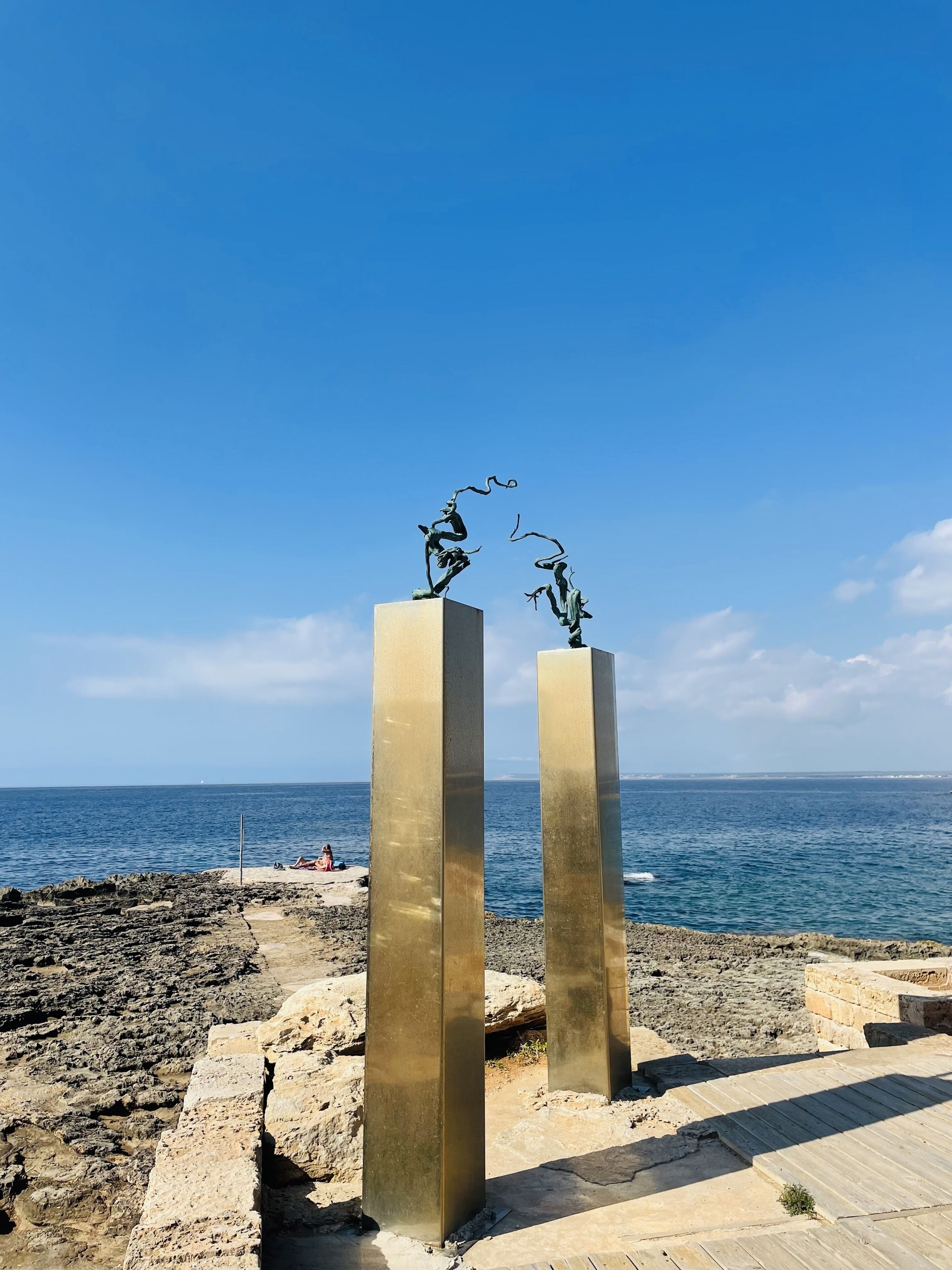 Two tall, rectangular bronze sculptures with abstract figures on top, set against a clear blue sky and the sea.