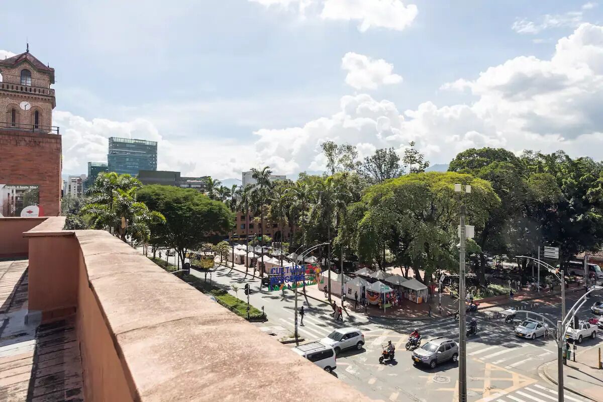 A busy urban street with a prominent clock tower, greenery, and modern buildings.