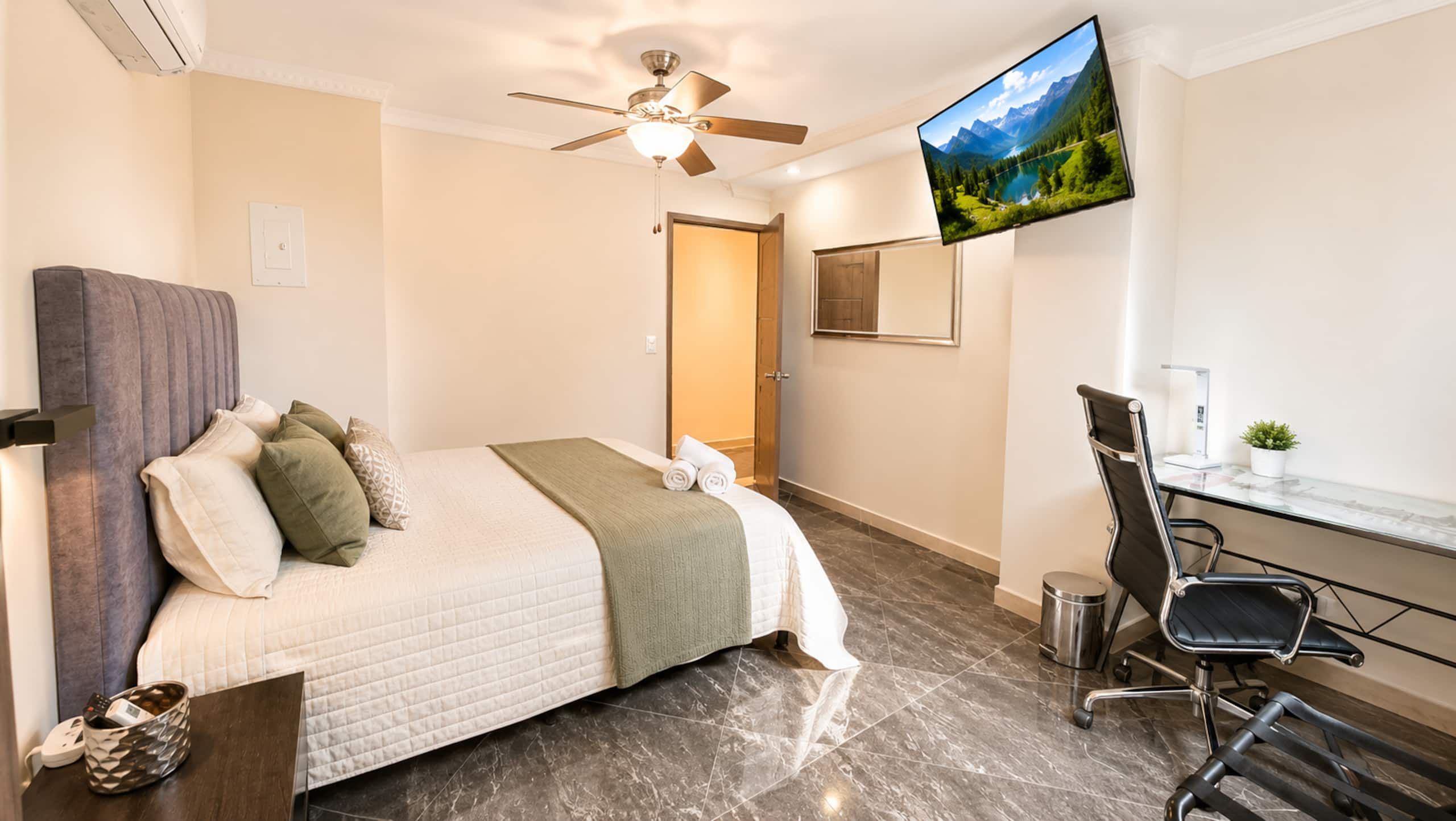 A hotel room with a bed, a desk, a chair, a TV, and a ceiling fan.
