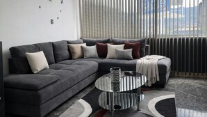 A modern living room with a grey sectional sofa, a glass coffee table, and a view of a cityscape through large windows with blinds. - Luxury Apartment! Amazing location! Poblado! Lleras! Provenza! Air Conditioning! (Antioquia)
