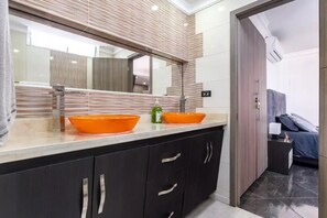Une salle de bain moderne avec un grand miroir, deux lavabos orange, des meubles sombres et une porte visible donnant sur une chambre.