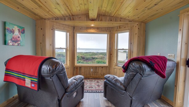 A room with two leather recliners, a large window with a view of the ocean, and a painting on the wall. - Sand Ranch - Epic Ocean View and SPA (Gold Beach)