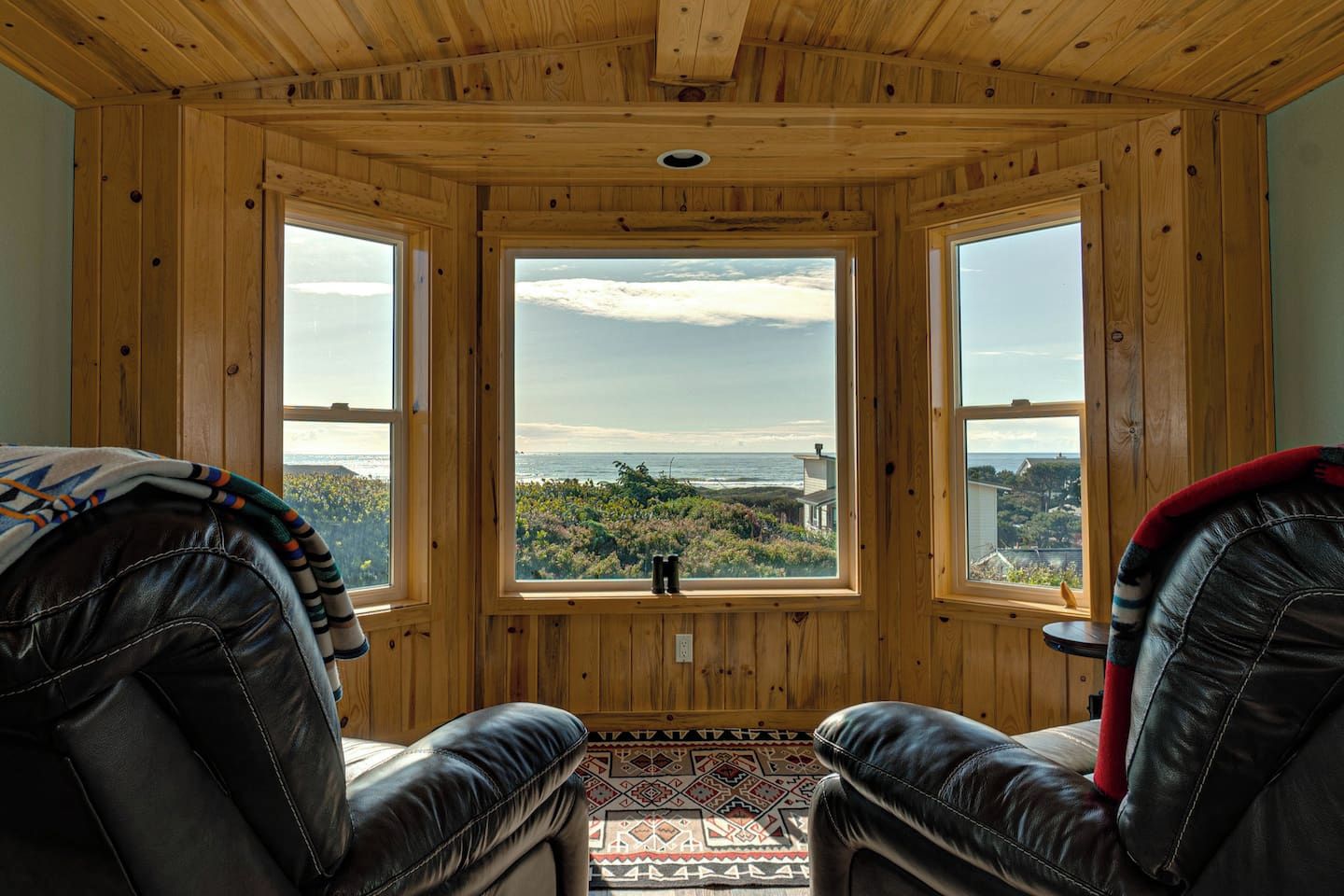 A cabin with a wooden interior, two leather chairs, a rug, and a view of the ocean and landscape through large windows.