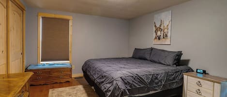 Quarto com cama, mesa de cabeceira, cômoda, janela com persianas e relógio de parede.