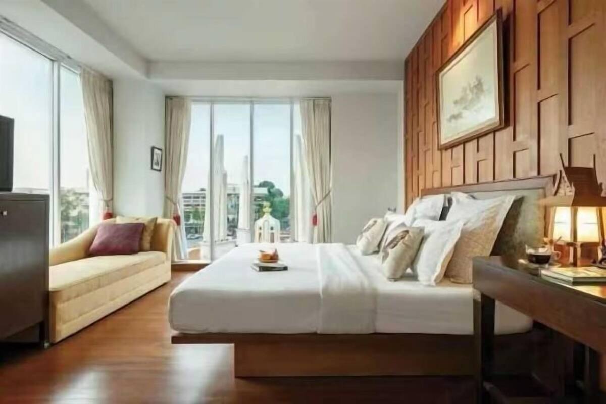 A hotel room with a bed, a sofa, a desk with a lamp, and a large window with curtains.