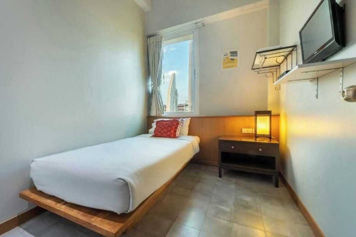 A single bed with a wooden platform, a bedside table with a lamp, a TV mounted on the wall, and a window with curtains.