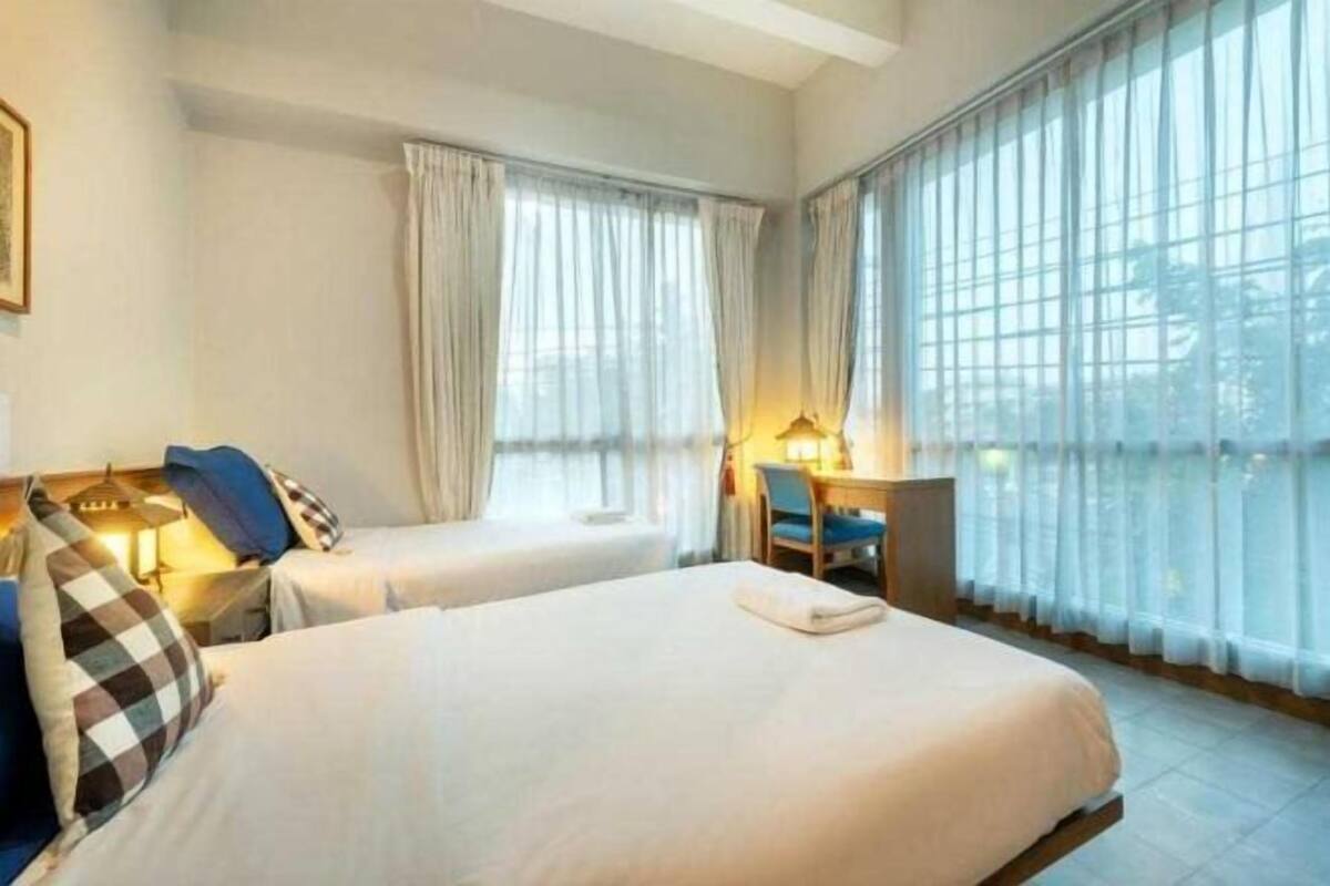 A hotel room with two beds, a desk, a chair, and large windows with curtains.