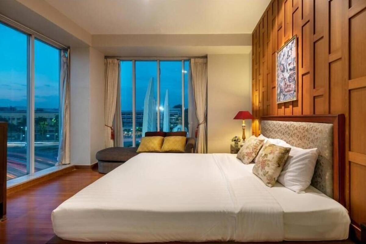 A large bed with a headboard, a nightstand, a lamp, and a view of a cityscape through large windows.