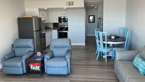 Ein modernes Wohnzimmer mit grauen Sofas, einem Esstisch mit blauen Stühlen und einer Küche im Hintergrund.