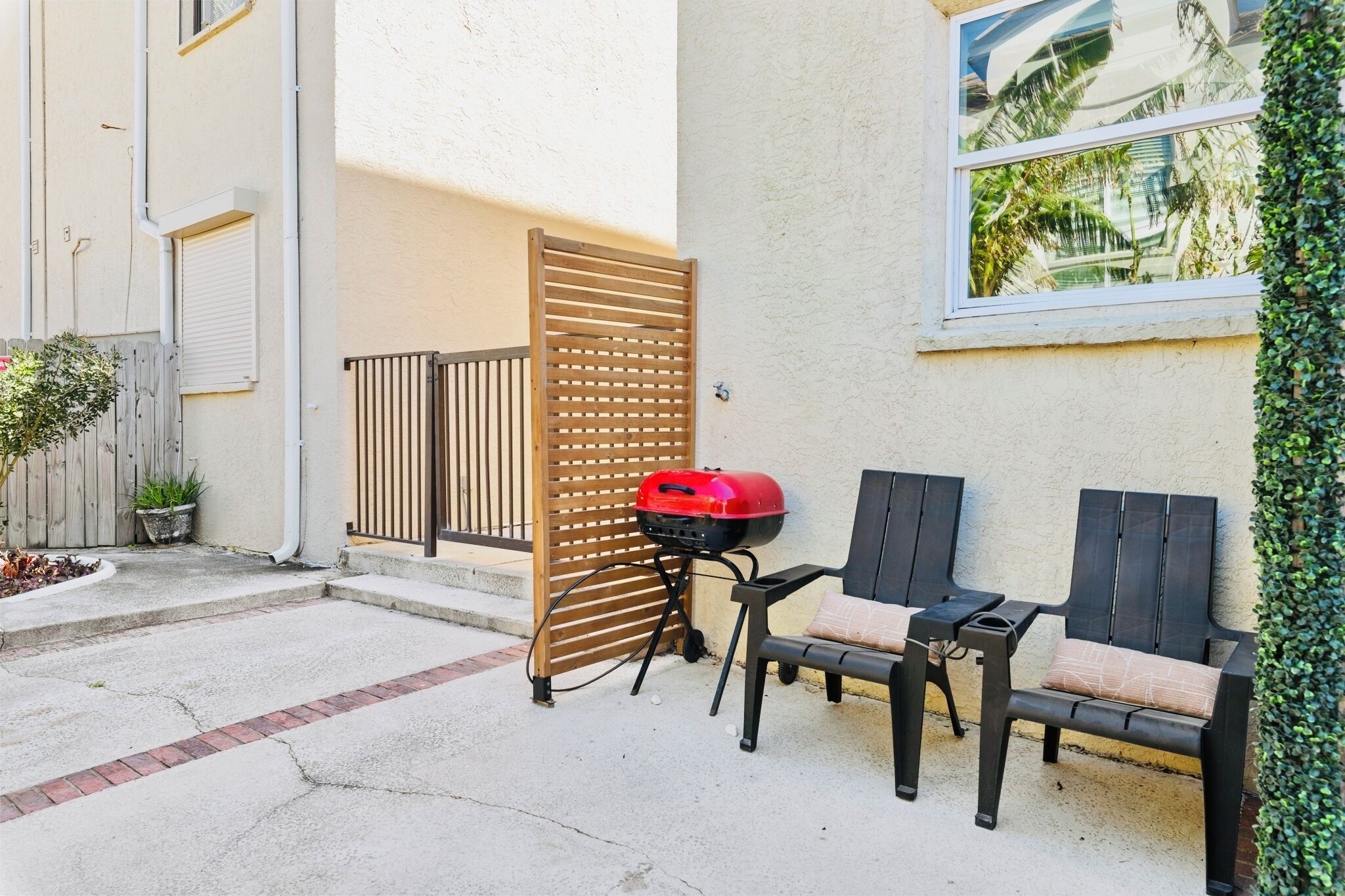 A patio with a grill, two chairs, a small table, a wooden gate, and a potted plant.