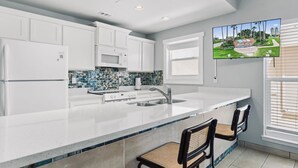 A modern kitchen with white cabinets, a large island with a sink, a microwave, and a stove. There are two bar stools and a television mounted on the wall.