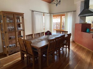 Un comedor con una mesa y sillas de madera, un aparador con cristalería y vistas a través de una ventana con cortinas.