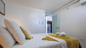 Ein Bett mit weißer Bettwäsche und einer gelben Decke, ein Nachttisch mit Handtüchern sowie eine Tür zu einer Küche.