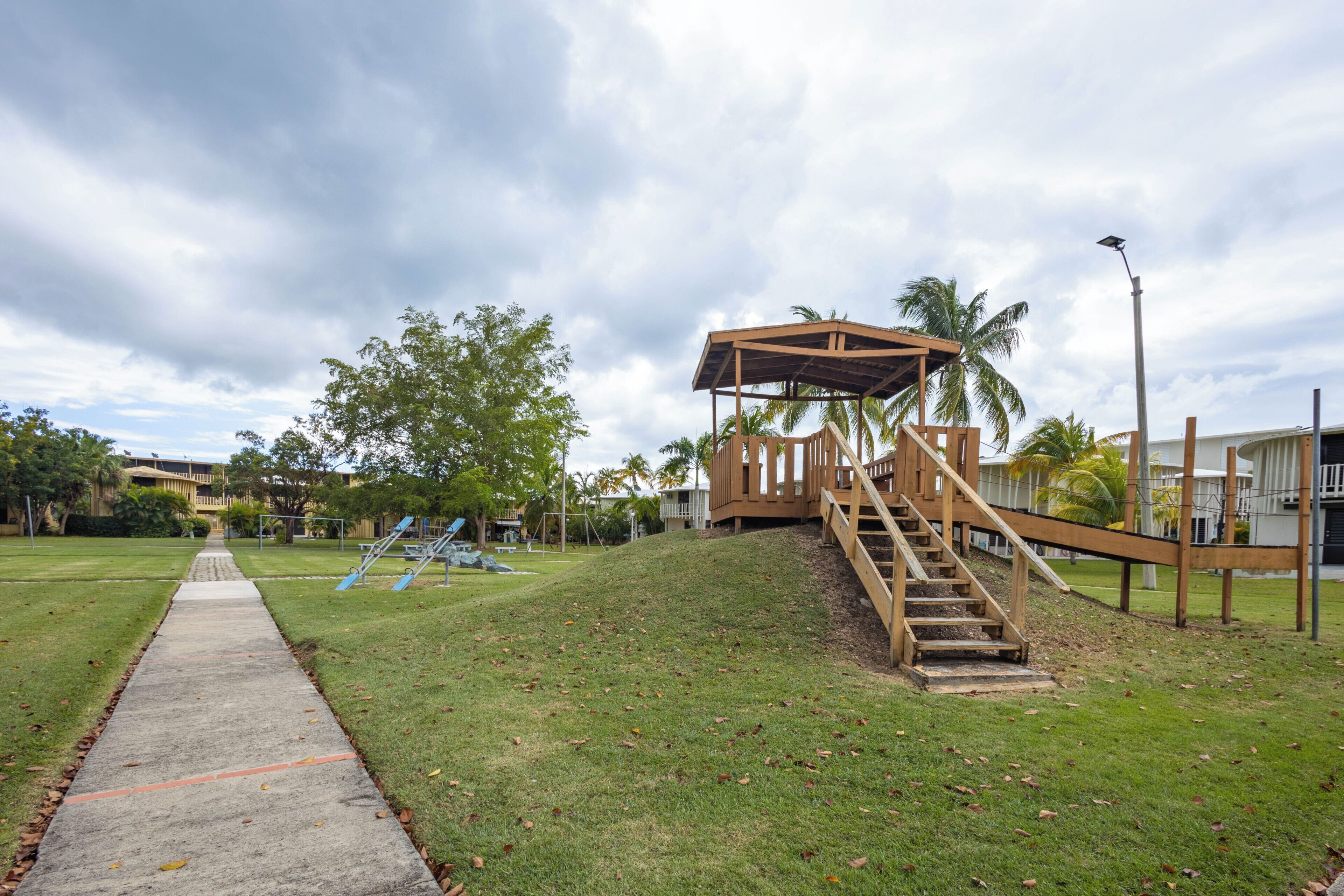 A park with a concrete path, a wooden play structure, and a staircase leading to a covered area.