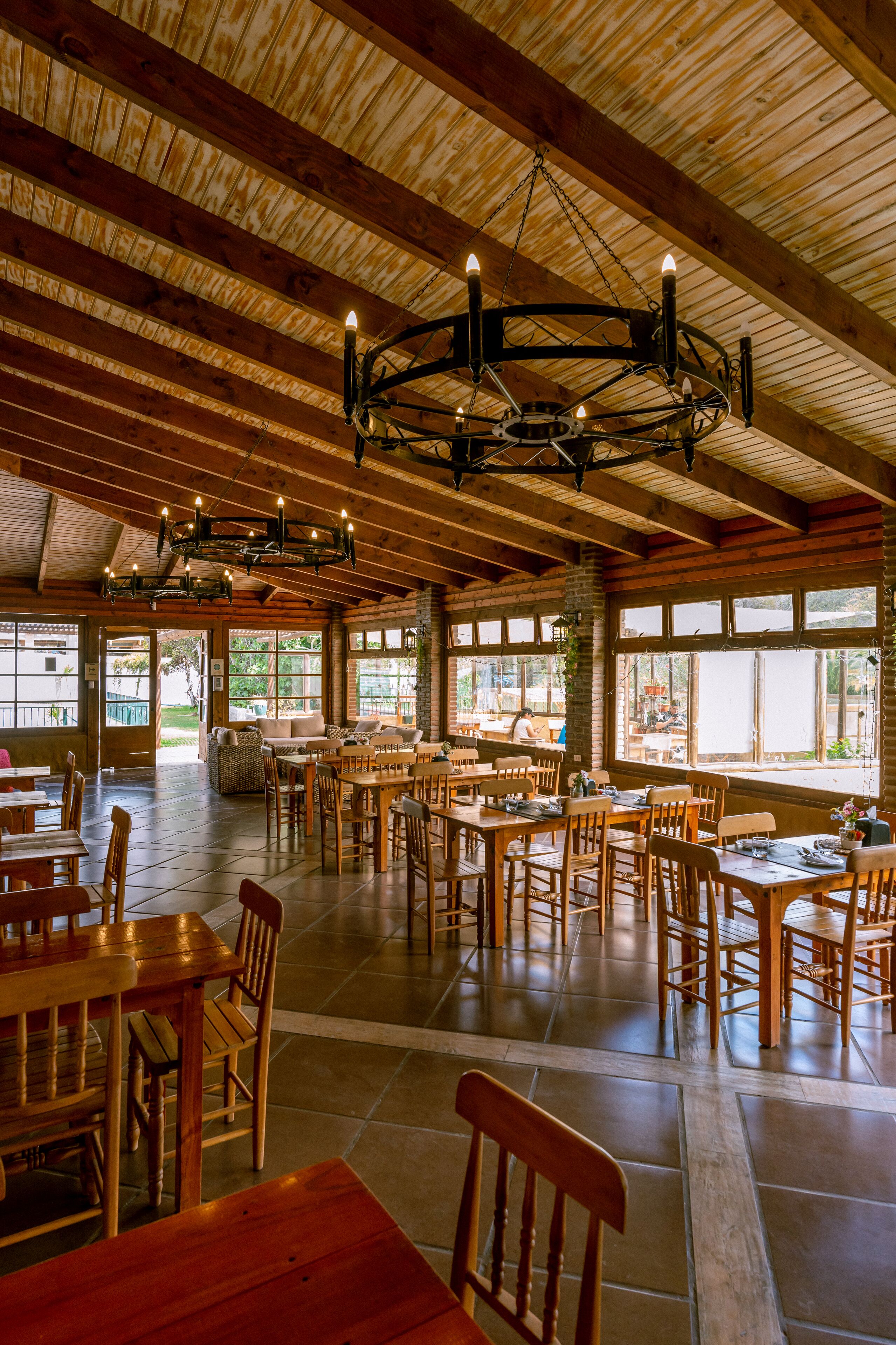 Un vaste espace dédié à la salle à manger, avec des tables et des chaises en bois, de grandes fenêtres et un plafond haut avec des poutres apparentes.