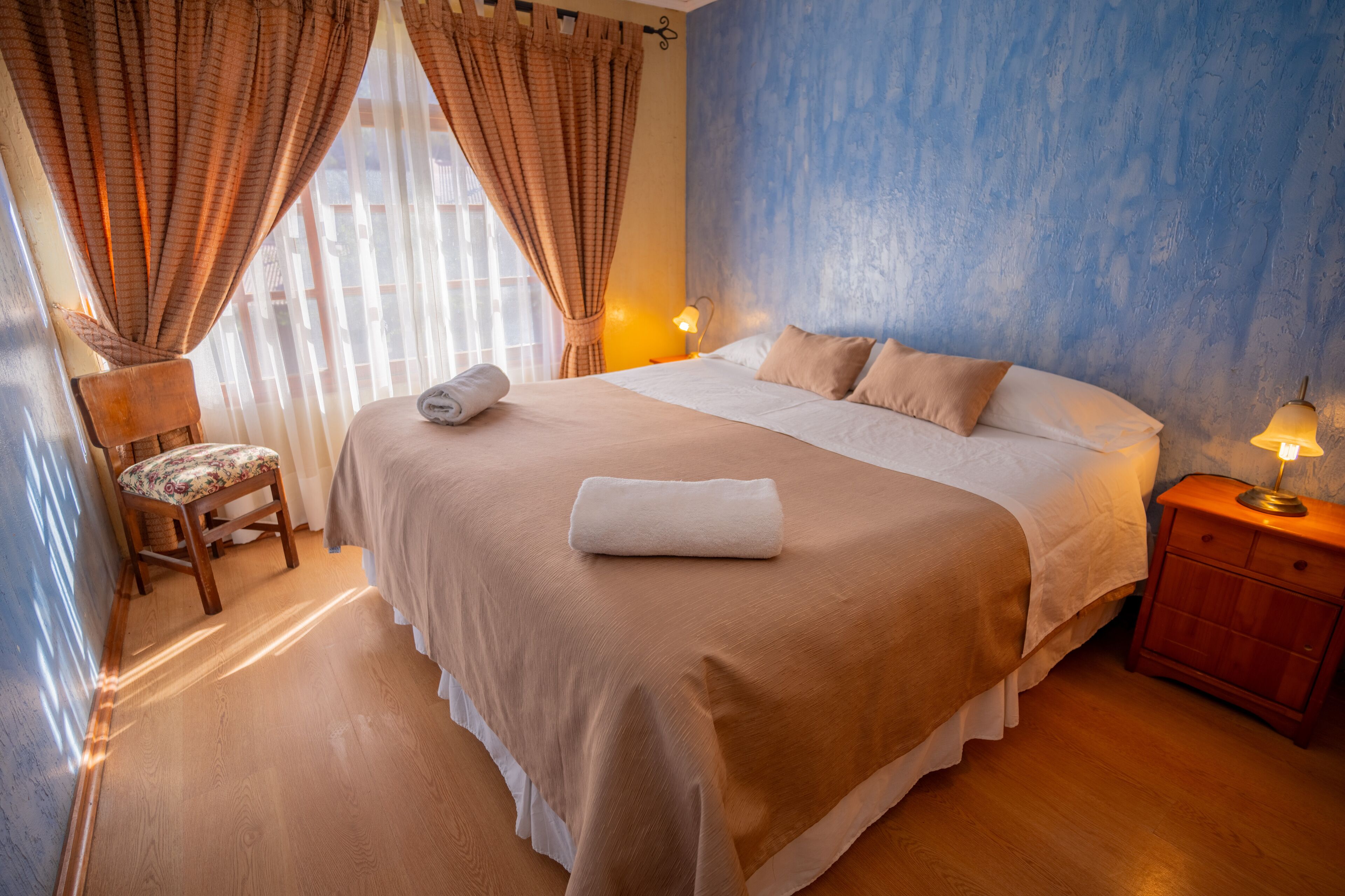 Una cama bien hecha con colcha blanca y beige, una mesita de noche de madera con una lámpara, y una silla con cojín estampado de flores.