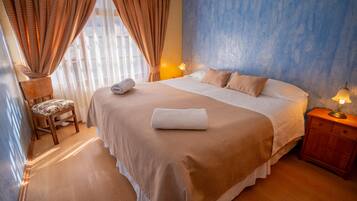 Una cama bien hecha con colcha blanca y beige, una mesita de noche de madera con una lámpara, y una silla con cojín estampado de flores.