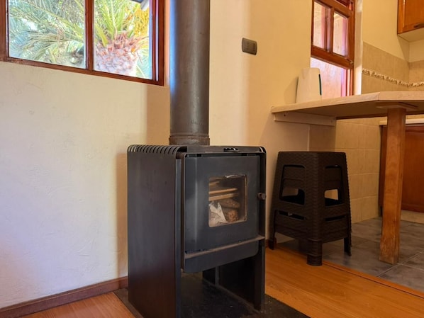 A wood-burning stove in a room with a window, a small stool, and a tiled floor.