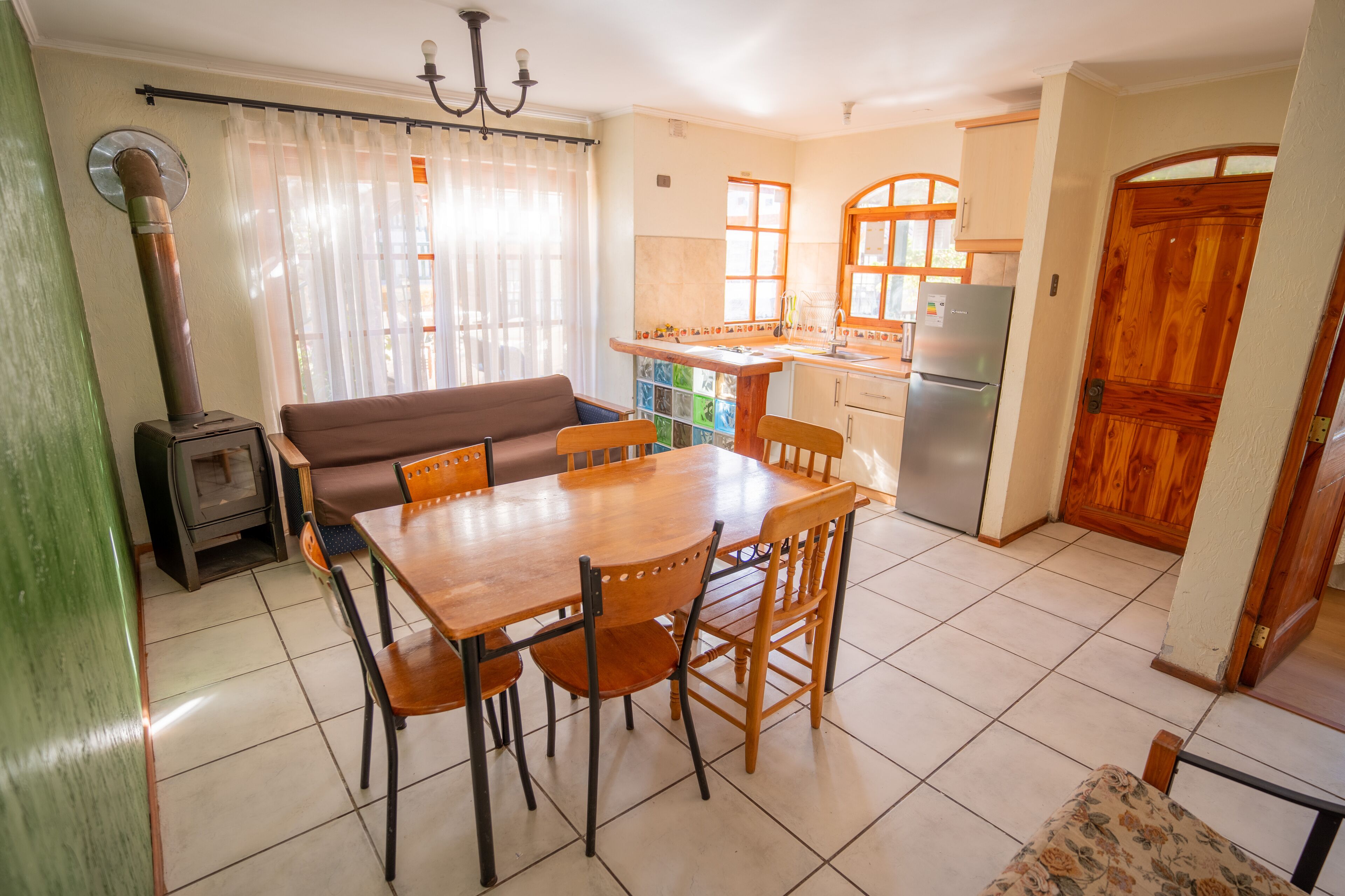 A dining area with a wooden table and chairs, a sofa, a kitchen with a refrigerator, and a wooden door.
