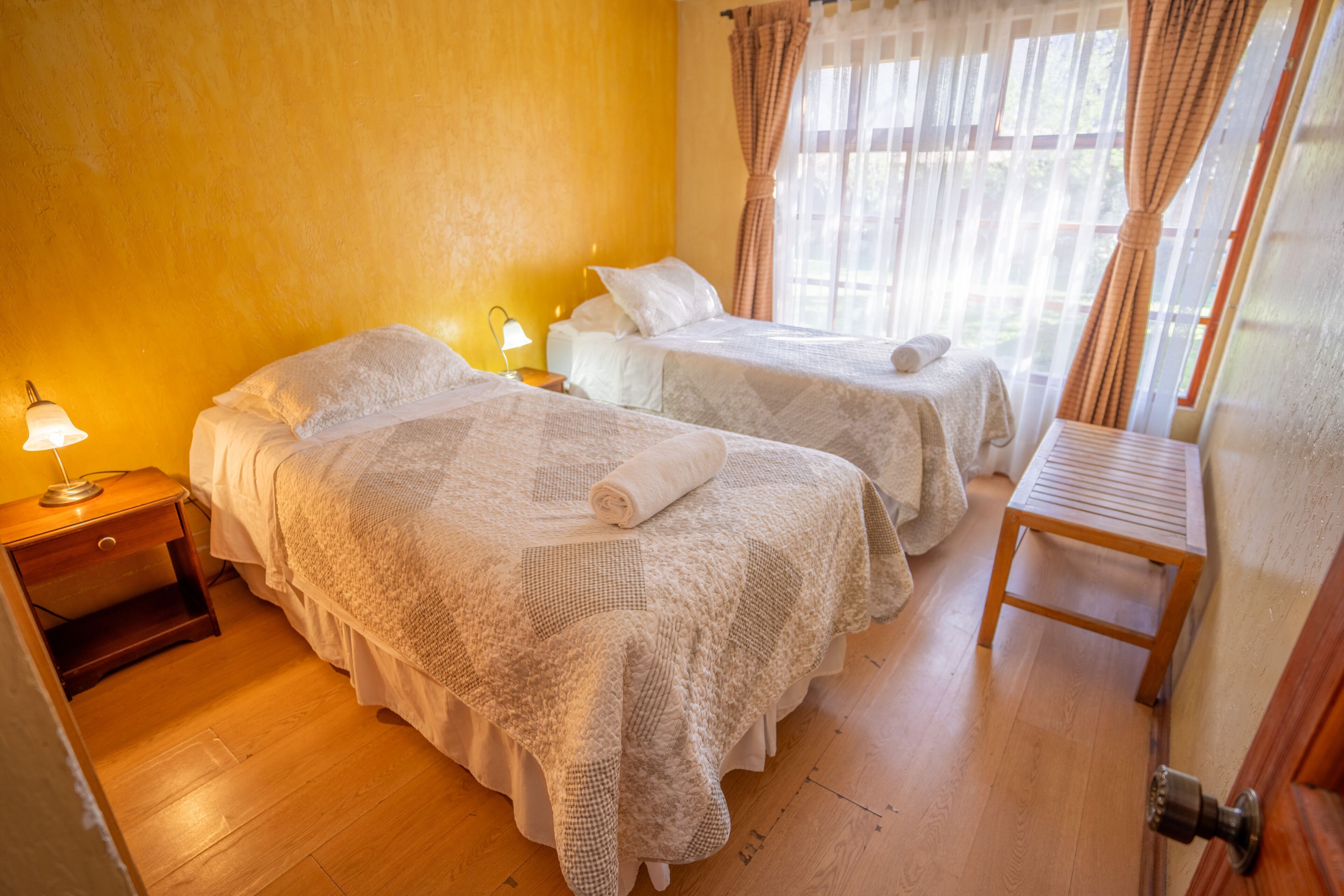 A hotel room with two beds, a wooden nightstand with a lamp, a wooden bench, and a window with curtains.