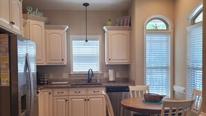 A kitchen with white cabinets, a stainless steel refrigerator, a dining table with chairs, and a window with blinds.