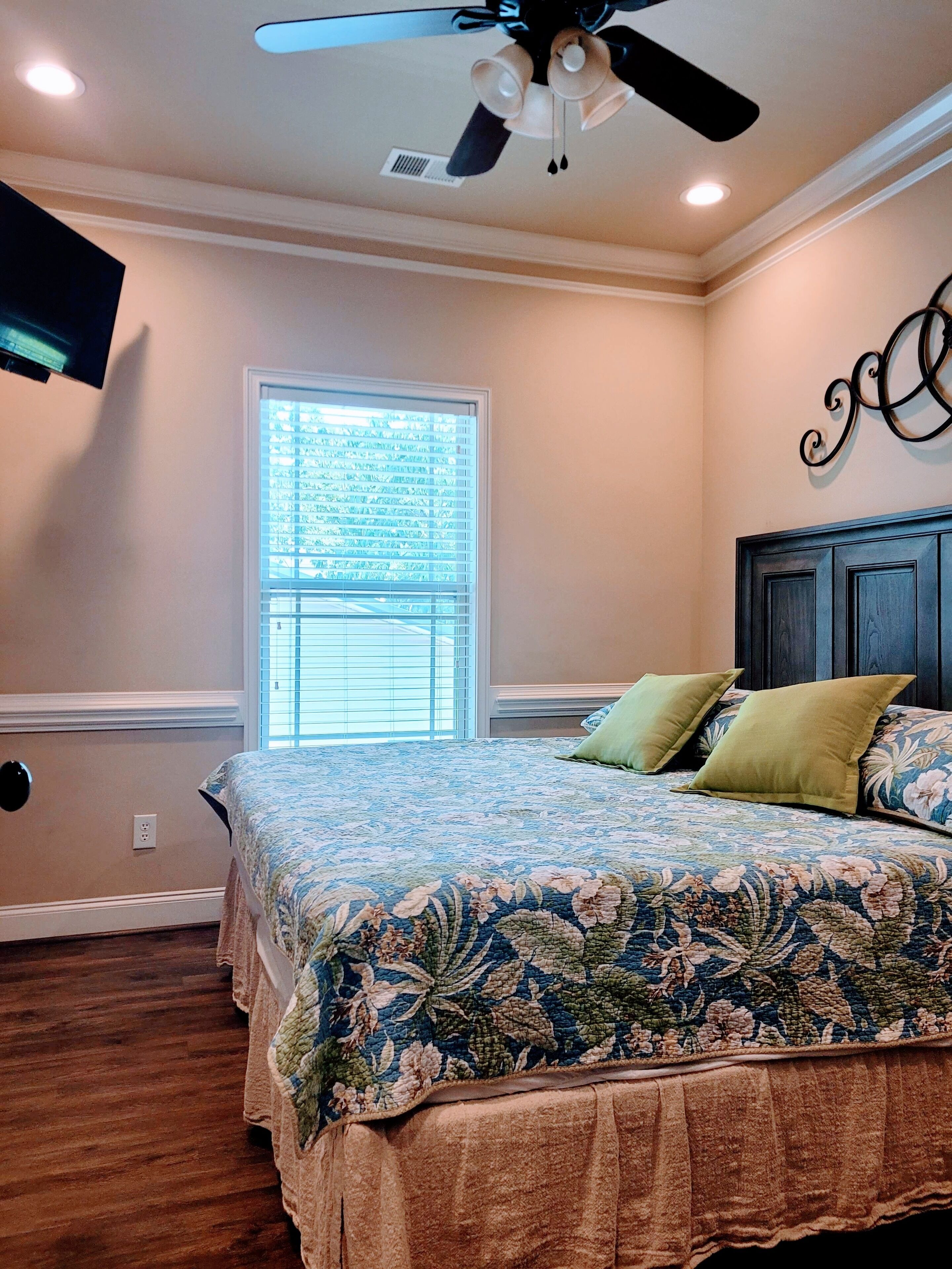 A bedroom with a bed, a ceiling fan, a window, and a wall-mounted TV.