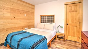 Un dormitorio con pared de madera, una cama con manta azul, un tapiz de cuadros y una puerta de madera, además de una mesita de noche con una lámpara.